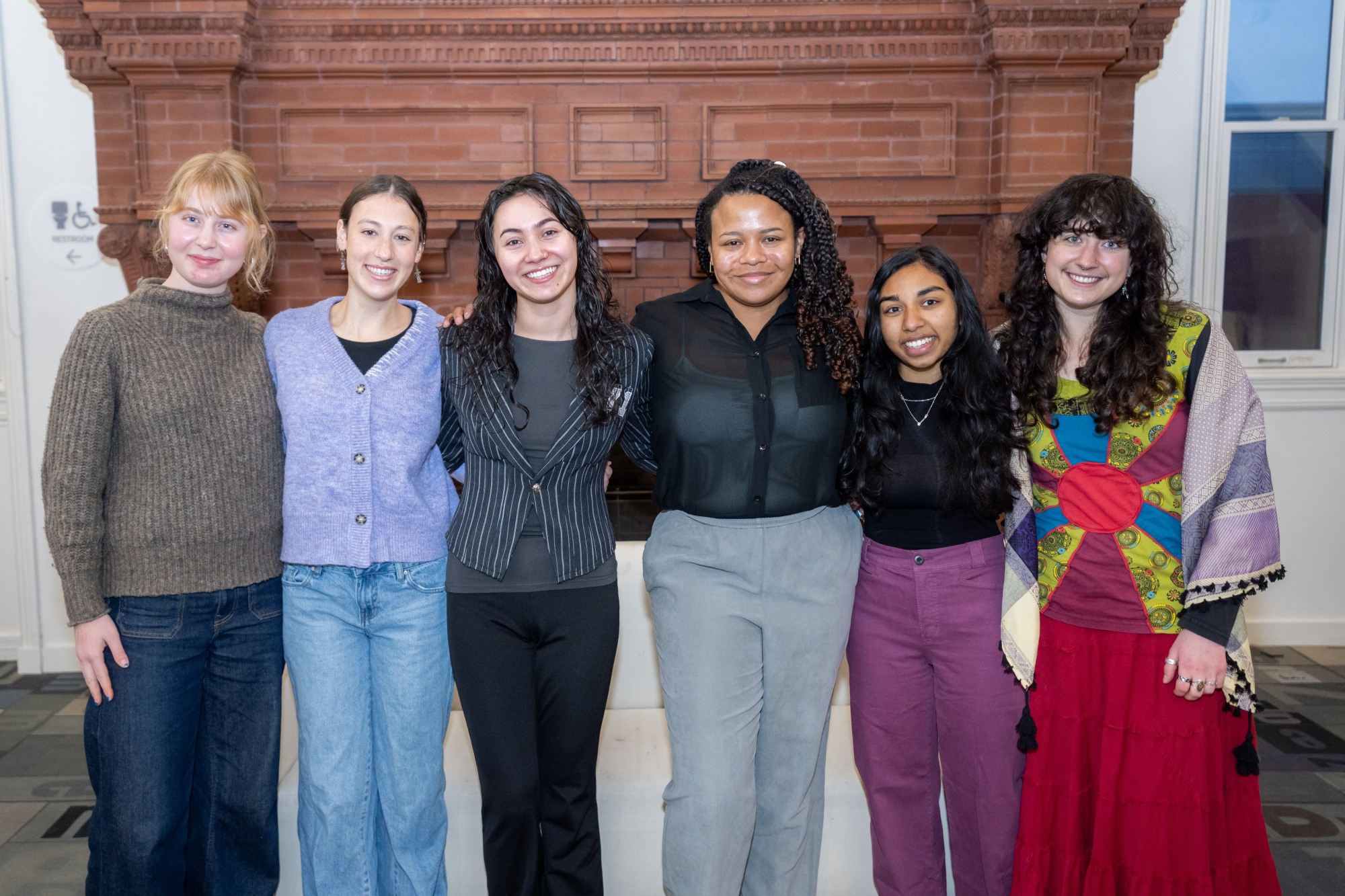 A group of six students stands in front of a large fireplace.