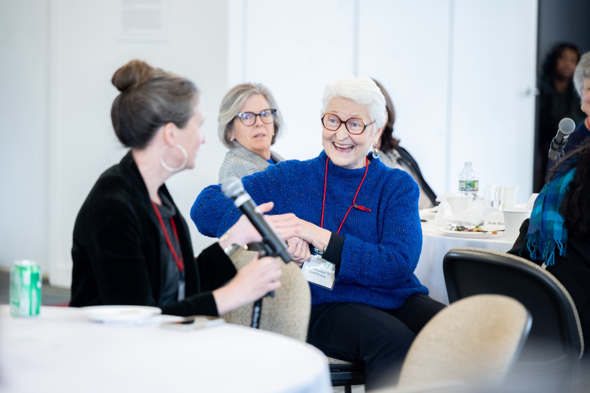 People sit at circular tables. Mary Murphy speaks into a microphone and Jean Howard reacts positively to what she is saying.