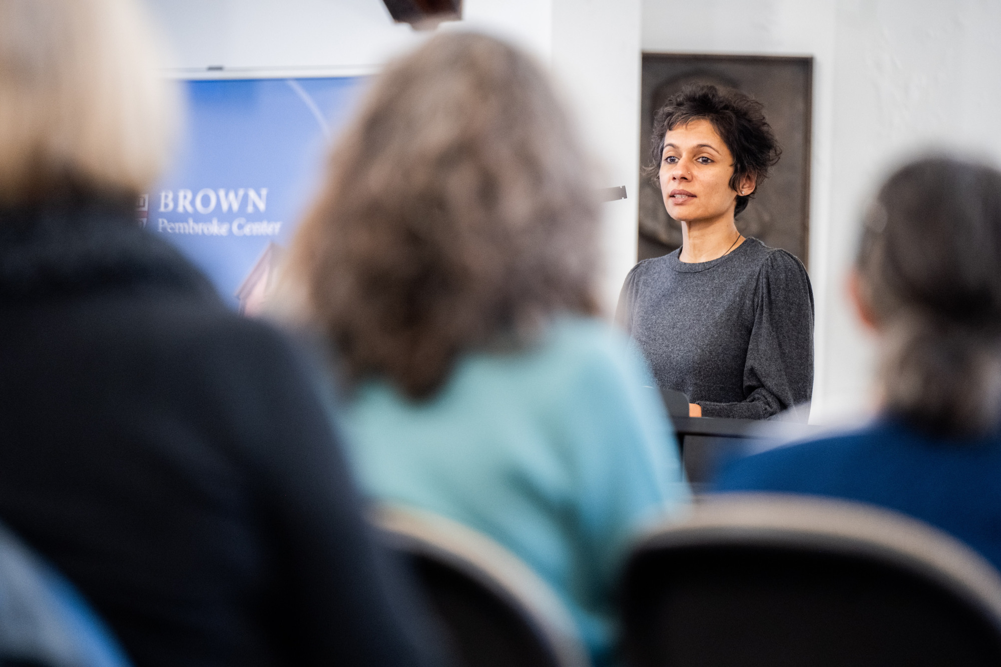 Thangam Ravindranathan speaks into a microphone at a lectern. Audience members' heads are in the foreground.