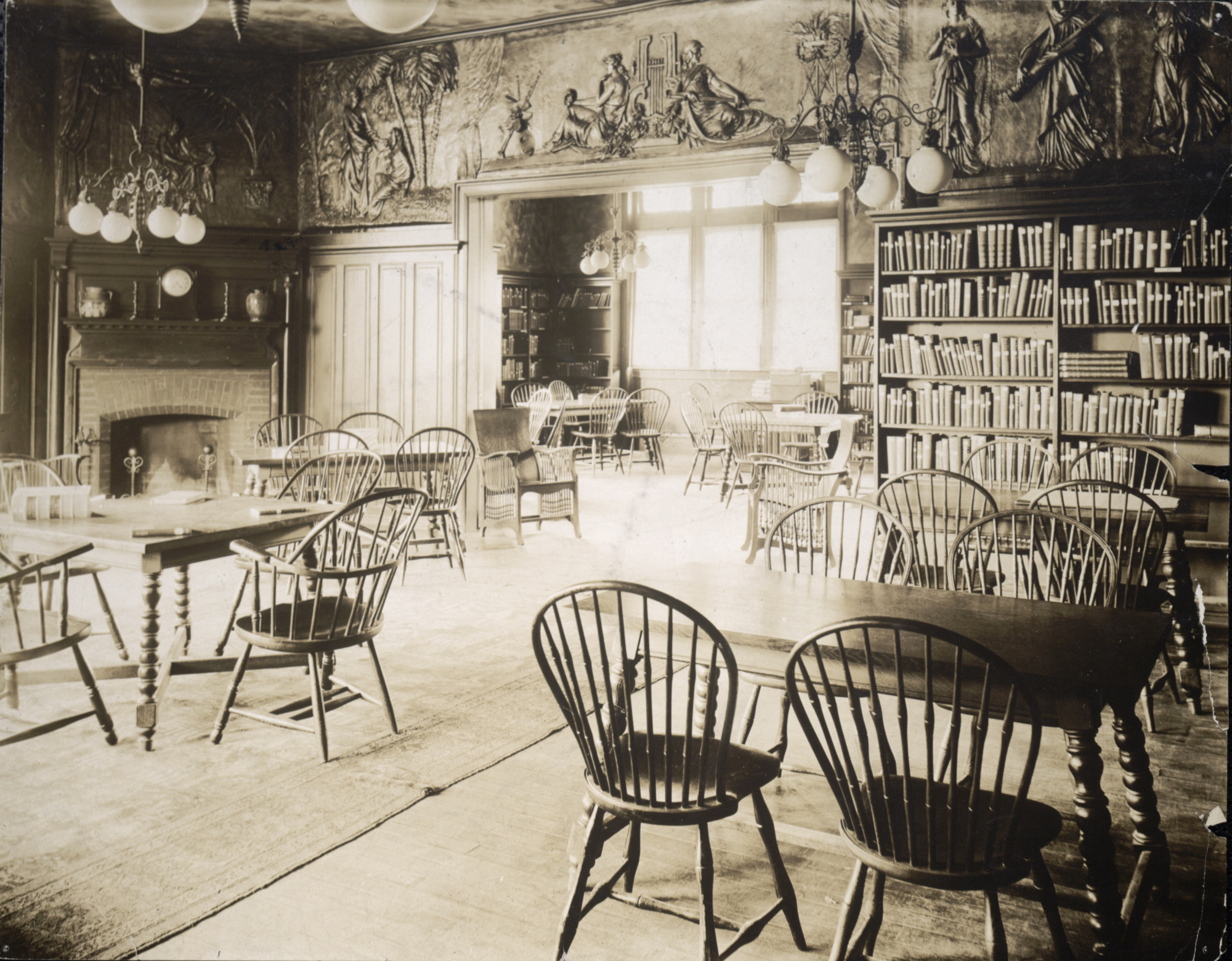Black and white image shows spacious, old-fashioned rooms with bookshelves along the walls and reading tables with chairs grouped around them. Sunlight is coming in from the windows.