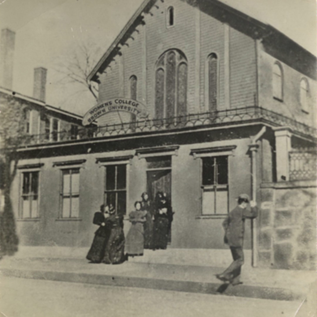 Black and white photograph showing a group of women standing at the entrance to the Women's College building. A man walking by is tipping his hat