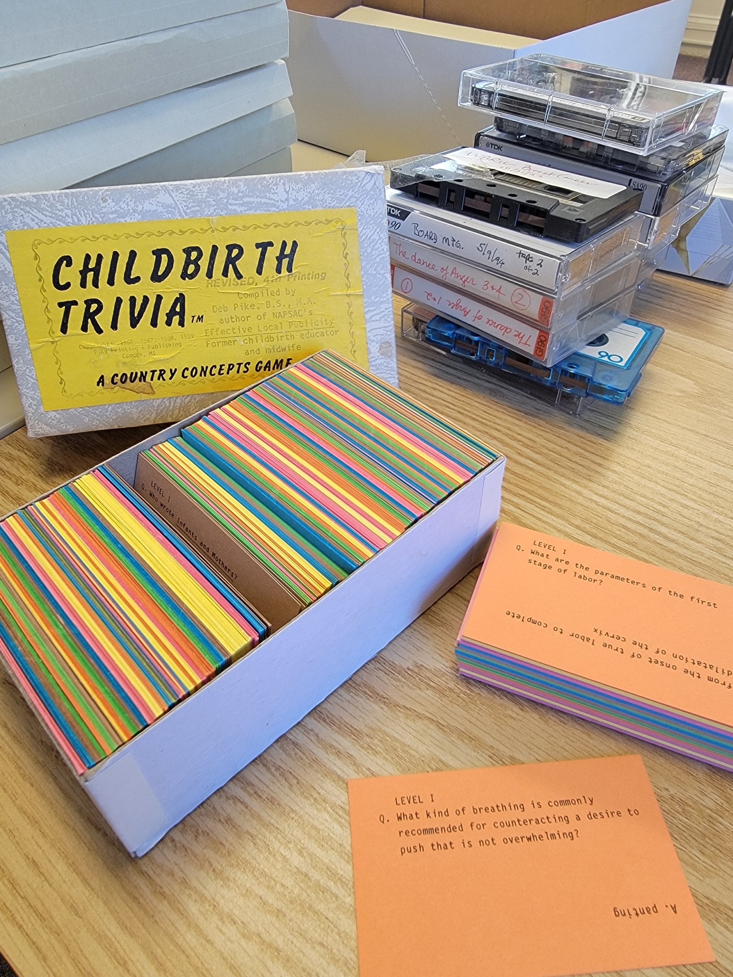 Image shows a box of index cards labeled "childbirth trivia." A pile of cassette tapes are behind the box, and other boxes of papers are behind it as well.