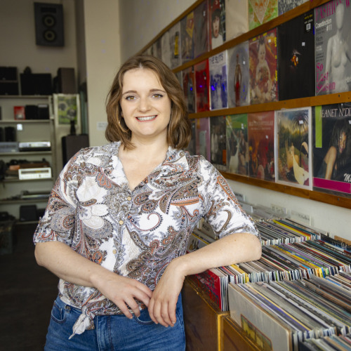 Headshot of Briana Nave, who is leaning against a shelf of records in a record shop.