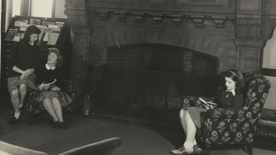 Black and white photograph shows three students sitting in armchairs by the large third floor fireplace.