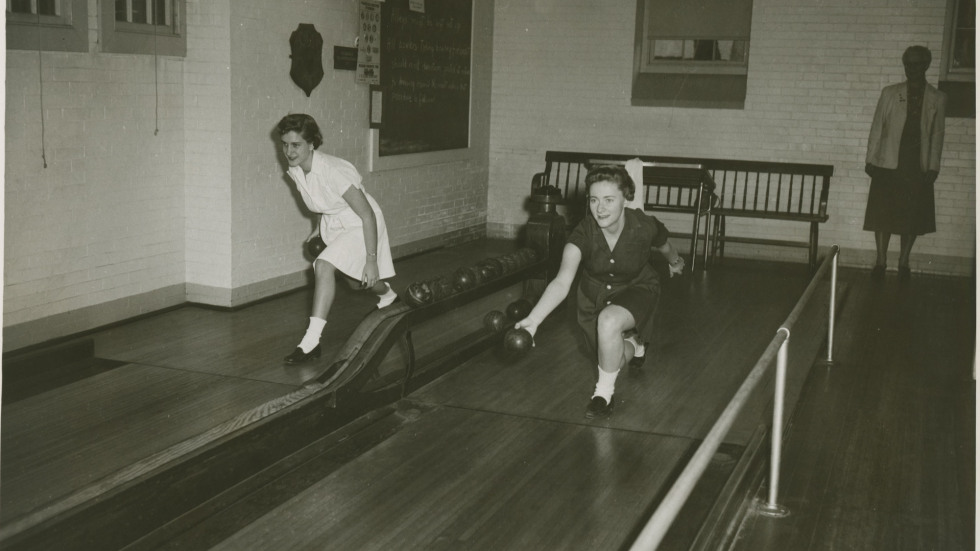 Black and white photograph shows two women bowling with a third watching. 