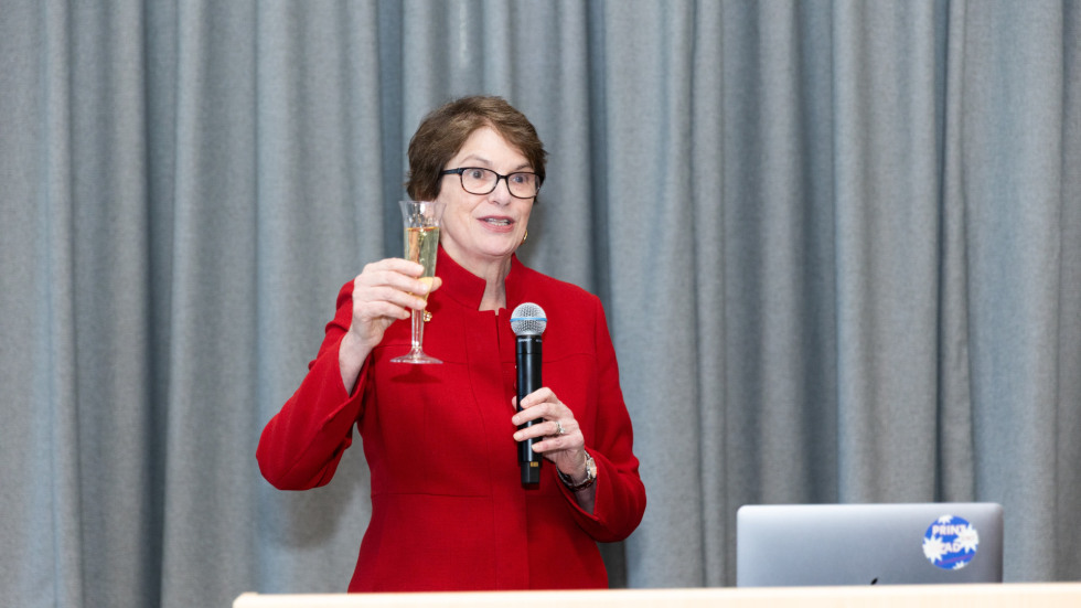 President Paxson stands at a lectern holding a microphone in one hand and a glass of champagne in the other.