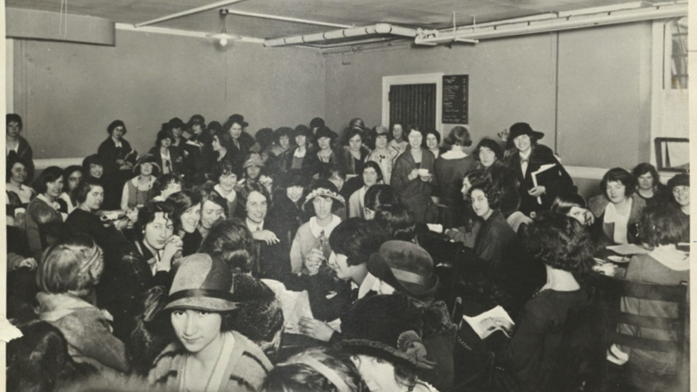 Black and white photograph shows women students crowded into a basement room seated, eating, and talking.