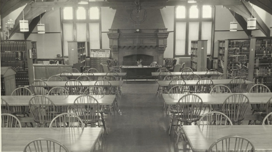 Black and white photograph shows room 305 in Pembroke Hall as the library. There are tables and chairs along the center, a fireplace in the middle of the far wall, and shelves to the left and right.