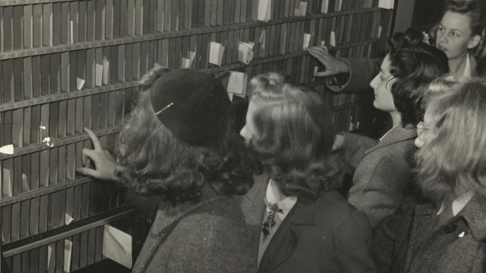 Black and white photograph of women students in hats and coats checking their mailboxes.